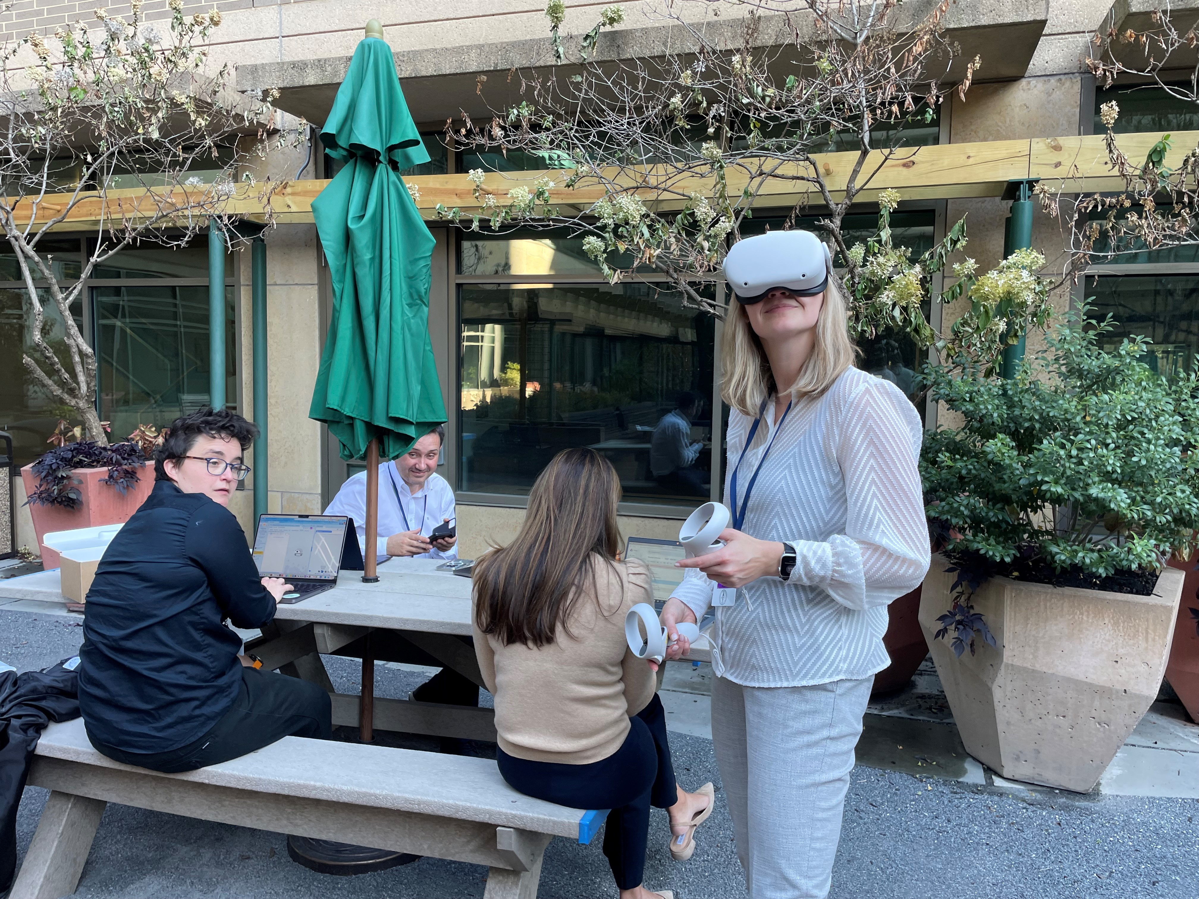 Several people gather around a concrete picnic table in an outdoor courtyard with planters and greenery. Some are seated and working on laptops while others stand nearby holding papers. The setting appears informal and collaborative, with a building and windows in the background.
